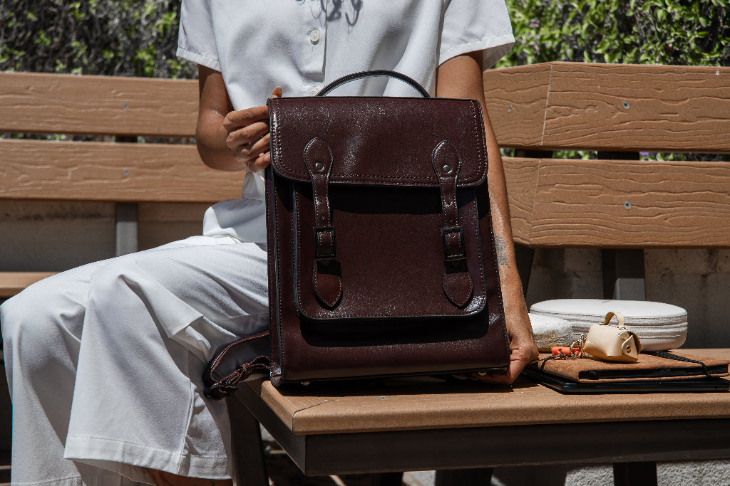 Person holding a brown leather backpack on a wooden bench outdoors.